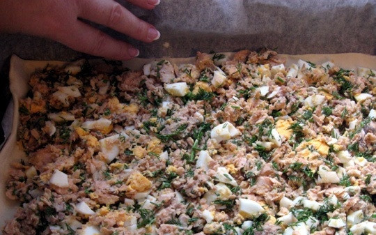 1. Этап. Пирог с красной рыбой из слоеного теста: Тесто разморозьте. Один пласт раскатайте и выложите на противень. Рыбу достаньте из банок, смешайте в отдельной посуде и помните вилкой. Добавьте к рыбе нарезанные и сваренные вкрутую яйца, рубленую зелень, соль, перец. Равномерно выложите рыбу на лист теста.