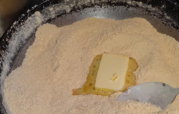 1. Этап. Армянская халва: Муку со сковороды не убираем! Пока она горячая, постепенно вводим масло, кусочками порезав его. Масло будет сразу таять, размешивайте его ложкой с мукой в тот же момент.