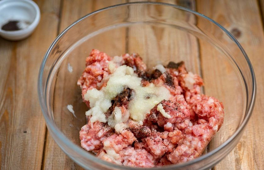 1. Этап. Зелёные пельмени со шпинатом: Приготовьте начинку для пельменей. В миску выложите фарш, натрите на мелкой терке очищенную луковицу, продавите через пресс очищенный зубчик чеснока, добавьте соль и черный молотый перец.