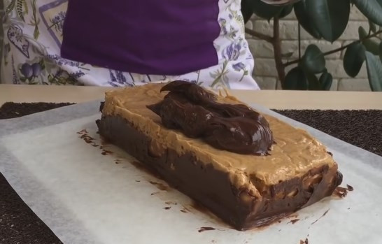 1. Этап. Настоящий торт Сникерс: 11. Извлеките торт из формы, отделите пергаментную бумагу. На новый лист пергамента выложите часть ганаша и распределите приблизительно по размеру торта. Положите на него батончик и ножом запечатайте края. Сверху тоже распределите ганаш ровным слоем.