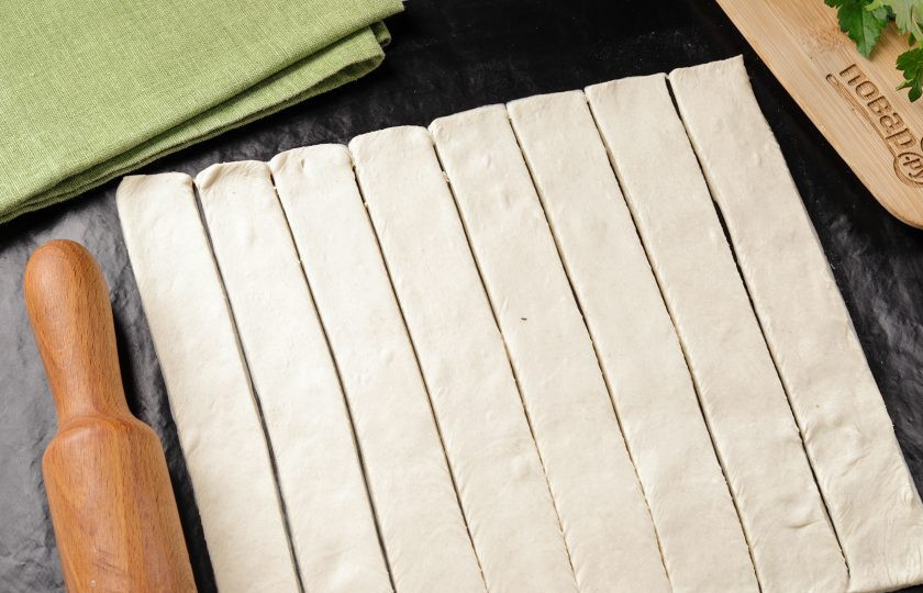 1. Этап. Сосиски в слоеном бездрожжевом тесте в духовке: Размороженный пласт теста раскатайте на слегка припыленном мукой столе, в прямоугольник толщиной около 3 мм. Размер готового прямоугольника будет зависеть от размера сосисок и их количества. Нарежьте прямоугольник на длинные полоски шириной около 1,5-2 см. по количеству сосисок. Длина полосок зависит от длины ваших сосисок.