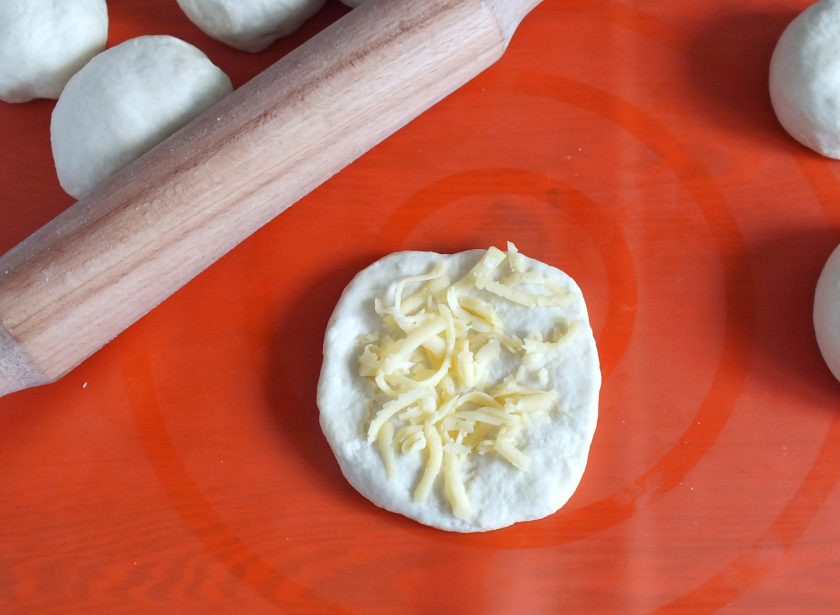 1. Этап. Булочки на рассоле: Возьмите заготовку из теста, раскатайте в лепешку. Сверху выложите пару щепоток тертого сыра.