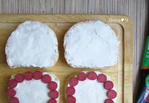 1. Этап. Бутерброд с колбасками и омлетом: Булочки намажьте плавленым сыром. Колбаски нарежьте тонкими кружочками и выложите по внешнему контуру булочки.