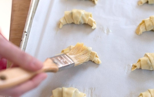 1. Этап. Мини круассаны: 3. Взбейте желток, добавив ложку водички. Смажьте с помощью кисточки поверхность круассанов и отправьте противень в разогретую духовку.