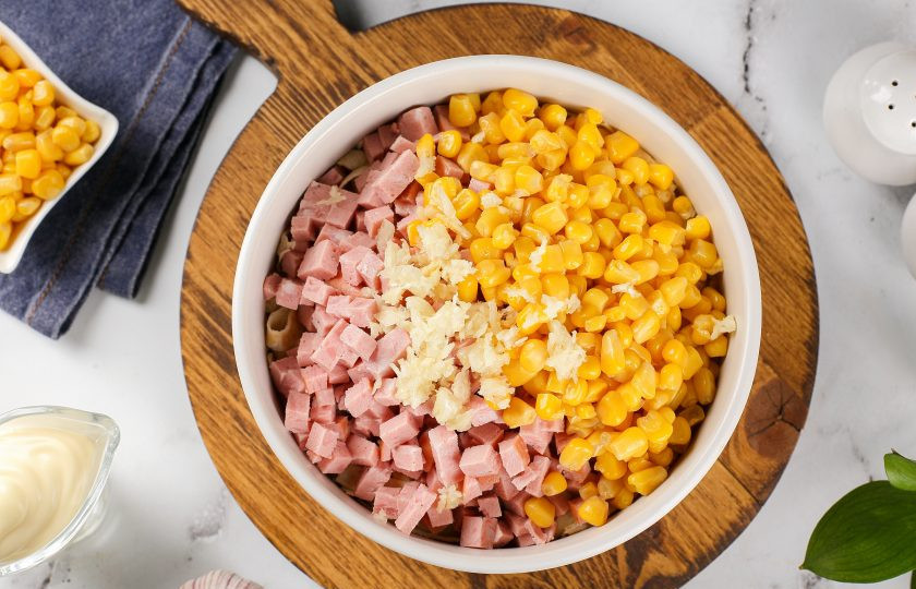 1. Этап. Салат с яичными блинчиками: Добавьте кукурузу, нарезанную кубиками колбасу и давленый чеснок.