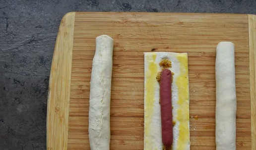 1. Этап. Колбаски Рилс в тесте: Лист теста разморозить и не раскатывая разрезать на три одинаковые части. По центру сделать чайной ложкой углубление и распределить по 1 ч. л. горчицы. Края теста смазать яичным желтком. По центру выложить колбаски.