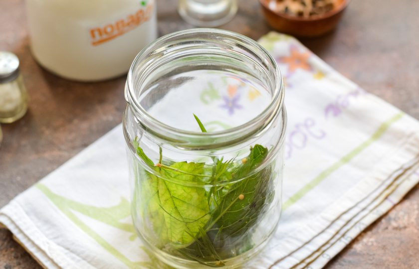 1. Этап. Огурцы с виноградными листьями на зиму: Банку для заготовки тщательно вымойте с пищевой содой, после ополосните хорошо под теплой проточной водой. Стерилизуйте банку и крышку удобным способом — над паром, в духовке или микроволновке. На дно банки положите все виды зелени, виноградные листья.