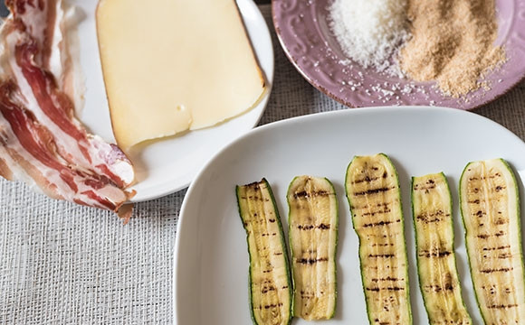 1. Этап. Закусочные рулетики из цуккини: 1. Вымойте цуккини, нарежьте их тонкими ломтиками и обжарьте на гриле по 4 минуты с каждой стороны, затем посолите и дайте им остыть. Приготовьте панировку из панировочных сухарей и тертого пармезана. Нарежьте бекон и сыр тонкими ломтиками.