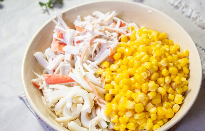 1. Этап. Салат с крабовыми палочками, кальмарами и кукурузой: Откройте консервированную кукурузу и слейте жидкость. Кукурузу добавьте к кальмарам и крабовым палочкам.
