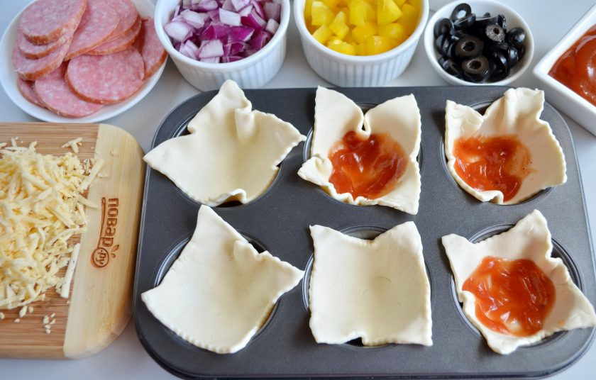 1. Этап. Закуска Пицца в стаканчиках: В форму для кексов вложите квадратики теста. Внутрь каждого квадратика положите по чайной ложке томатного соуса. Форма у меня антипригарная, поэтому я ничем не смазывала.