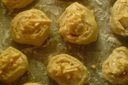 1. Этап. Булочки с сыром из слоеного теста: Сверните тесто в один рулет. Нарежьте небольшими ломтиками (примерно в один сантиметр). Выложите на пергамент и посыпьте мукой. Далее булочки смажьте яйцом. Для аромата можно положить пару долек чеснока и досыпать остатки сыра.