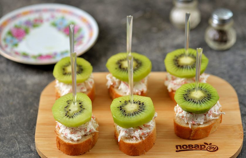 1. Этап. Бутерброды со сливочным сыром, киви и крабовыми палочками: Сверху на каждый бутерброд выложите по ломтику киви, скрепите все шпажкой или зубочисткой.