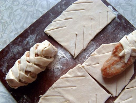 1. Этап. Фарш в тесте: Тесто заранее размораживаем, но не до конца, а чтобы разделить на отдельные листы. Раскатываем тесто (слоеное тесто раскатывают в одном направлении), нарезаем его на прямоугольники, а затем надрезаем с каждой стороны по диагонали елочкой. Кладем на прямоугольник котлету и начинаем заплетать косичку, накладывая полоски внахлест поочередно.