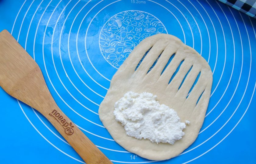 1. Этап. Отрывной пирог из дрожжевого теста с творожной начинкой: После достаньте его из миски и обомните. Разделите на 7 частей. Каждую скатайте в шарик. Творог перемешайте с сахаром до однородной консистенции. Шесть шариков раскатайте в овал. На одном крае сделайте надрезы. А на другой край выложите творожную начинку.