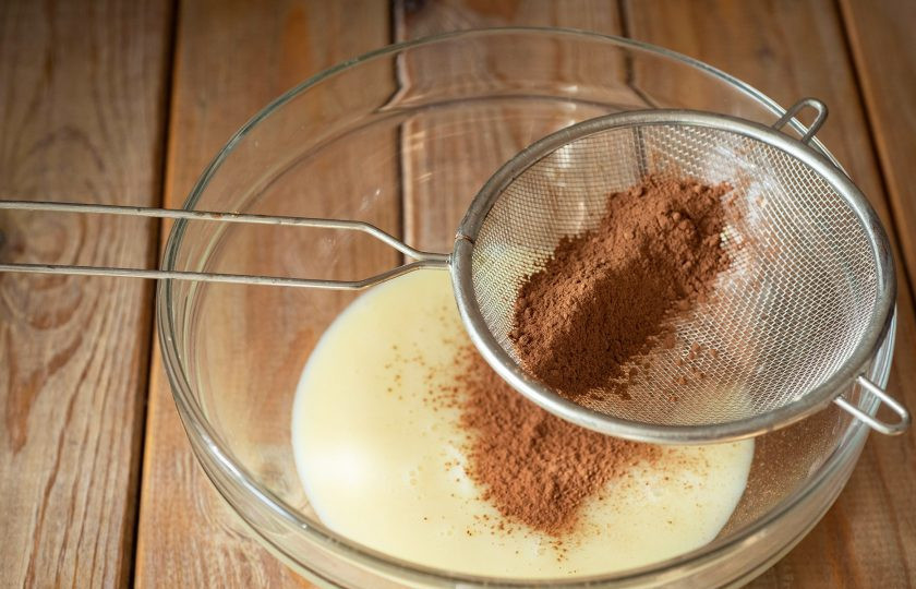 1. Этап. Конфеты из сгущённого молока и какао: Для удобства горячую сгущенку перелейте в просторную миску. Частями просеивайте к ней какао.