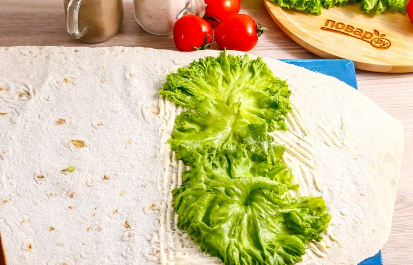 1. Этап. Курица в лаваше: Разверните на рабочей поверхности лист лаваша, обмажьте его 1 ст.л. майонеза и выложите на соус промытые салатные листья.