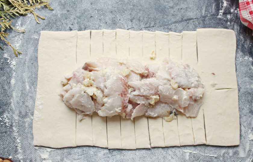 1. Этап. Пирог с муксуном: Стол посыпьте мукой и немного раскатайте. На середину выложите начинку. Разрежьте тесто с двух сторон на полоски шириной два сантиметра, не доходя с двух сторон примерно 4-5 сантиметров.