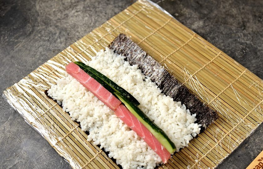 1. Этап. Роллы с тунцом и кунжутным соусом: Отрежьте от листа нори полоску толщиной 3-4 сантиметра, выложите слой риса, оставив противоположный край водорослей свободным. Выложите ломтики тунца и огурца на слой риса, скрутите ролл с помощью циновки.