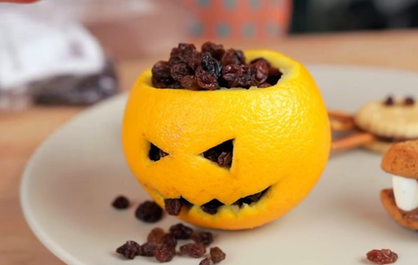 1. Этап. Снеки к Хэллоуину: 4. Далее приготовьте голову Джека. Для этого аккуратно срежьте верх с апельсина и с помощью ложки очистите его от средины. На кожуре с помощью канцелярского ножа вырежьте глаза и рот Джека, затем насыпьте в средину изюм. Прикройте крышкой.