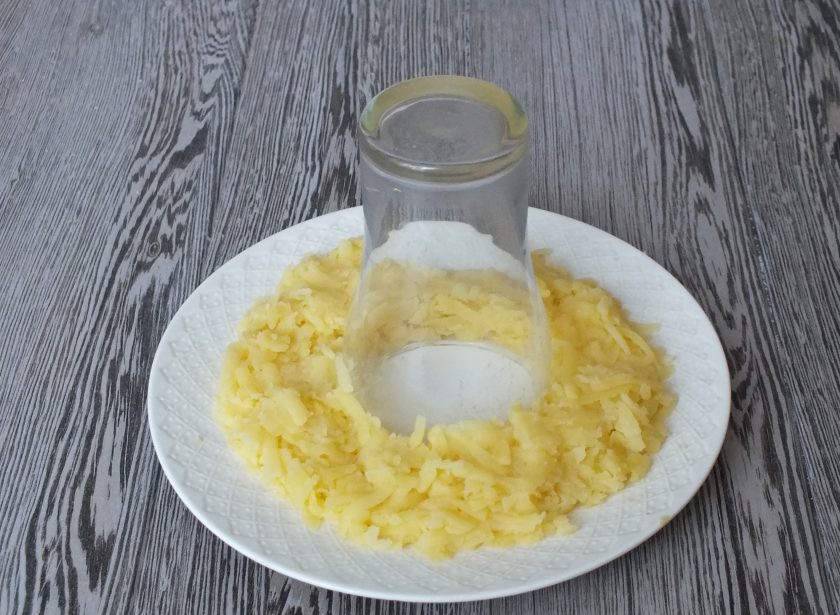 1. Этап. Салат Гранатовый браслет с черносливом и грецким орехом: Возьмите плоское блюдо и стакан. Картофель натрите на терке. По середине блюда поставьте стакан горлышком вниз. Вокруг стакана разложите тертый картофель, подсолите.