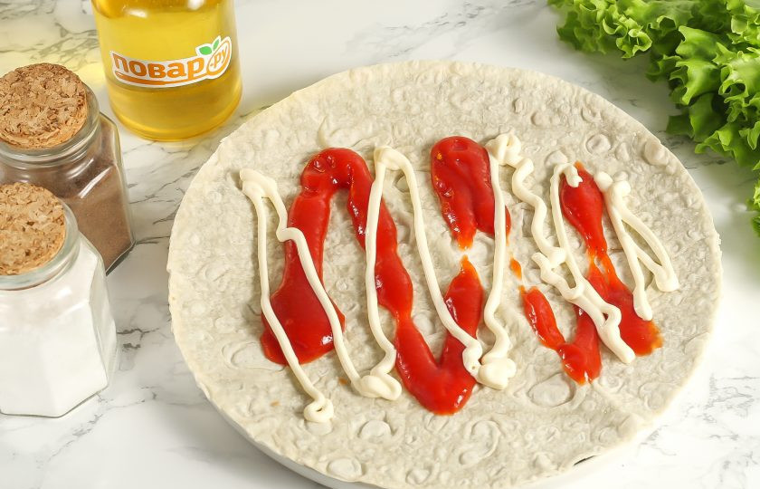 1. Этап. Цезарь ролл с наггетсами: Лепешку тортильи или круглый лист лаваша выложите на тарелку и обмажьте кетчупом и майонезом. Вместо майонеза можно использовать чесночный соус, если готовите ролл для взрослых.