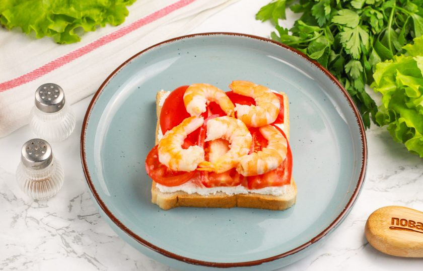 1. Этап. Сэндвич с креветками: Очищенные хвосты креветок выложите на помидоры. По желанию можно нарезать их кубиком или слайсами. Посолите креветки, по желанию — поперчите.
