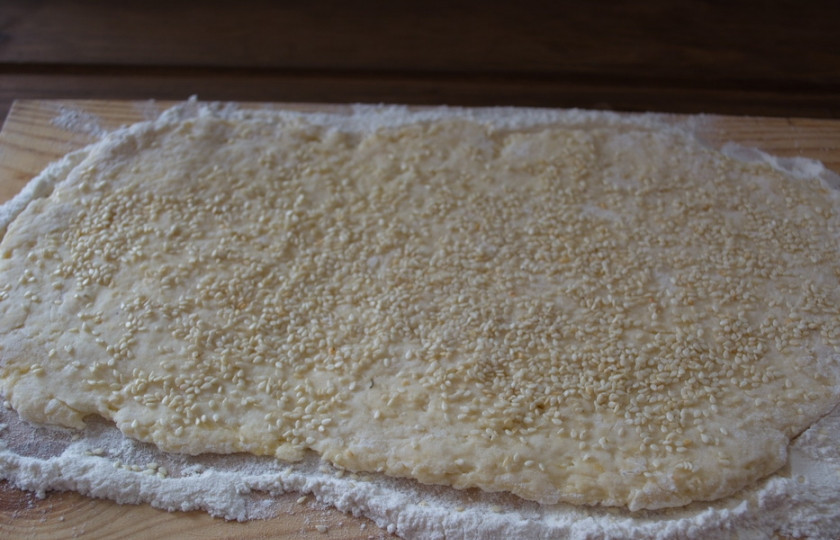 1. Этап. Печенье на сметане с кунжутом: Хорошо присыпьте доску мукой. Раскатайте тесто на доске. Сверху притрусите кунжутом и похлопывающими движениями закрепите кунжут на тесте, чтоб он не осыпался.