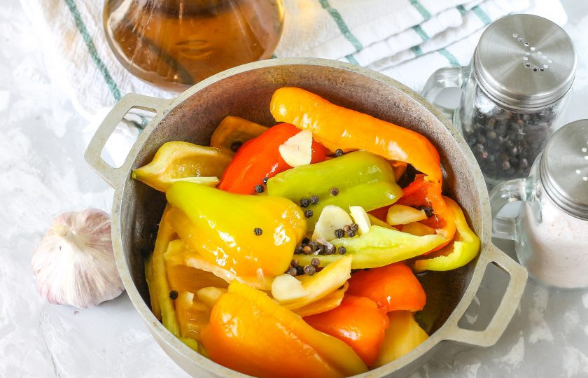 1. Этап. Перцы на зиму с медом: Выложите ломтики перца в казан, всыпьте горошины черного перца. Очистите и промойте чесночные зубчики, разрежьте их и добавьте в емкость.