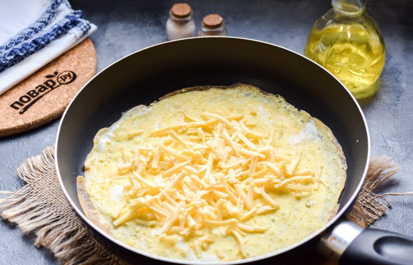 1. Этап. Рецепт завтрака из яиц: Сыр натрите на средней терке, выложите половину сыра на яичный блинчик.