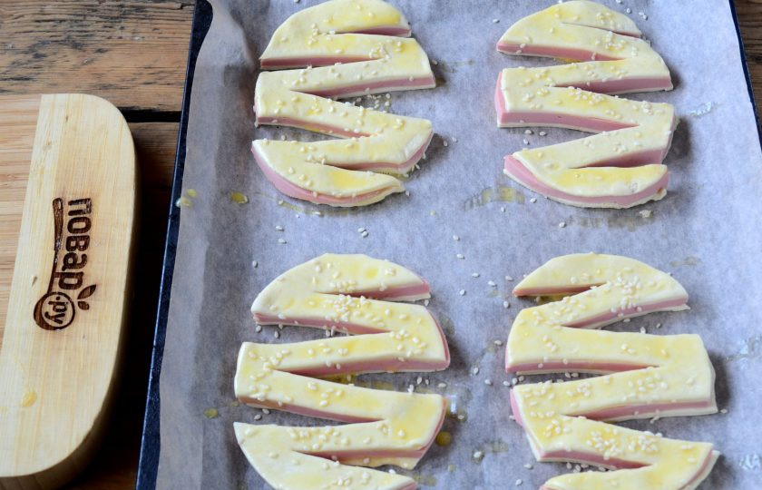 1. Этап. Слойки с колбасой Зигзаг: Переложите зигзаги на противень, застеленный пергаментом, смажьте взбитым яйцом и присыпьте кунжутом.