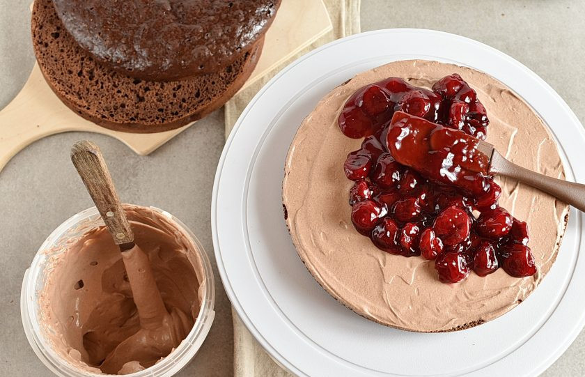 1. Этап. Торт в мультиварке: Смажьте нижний корж половиной сметанного крема. Выложите сверху охлажденную вишневую начинку.